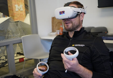A man wearing a "Concussion Center" branded pullover is seated indoors wearing a white virtual reality (VR) headset with a multicolored reflective sticker on the front labeled "VR Experience the Future." He is holding two white VR controllers, one in each hand, with his thumbs poised over the buttons, indicating he is actively engaged in a VR session.