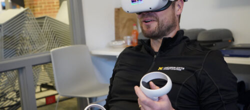 A man wearing a "Concussion Center" branded pullover is seated indoors wearing a white virtual reality (VR) headset with a multicolored reflective sticker on the front labeled "VR Experience the Future." He is holding two white VR controllers, one in each hand, with his thumbs poised over the buttons, indicating he is actively engaged in a VR session.