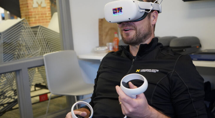 A man wearing a "Concussion Center" branded pullover is seated indoors wearing a white virtual reality (VR) headset with a multicolored reflective sticker on the front labeled "VR Experience the Future." He is holding two white VR controllers, one in each hand, with his thumbs poised over the buttons, indicating he is actively engaged in a VR session.