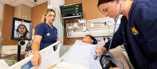 In a clinical simulation room, two nursing students, wearing U-M Flint uniforms, perform a medical checkup on a patient mannequin. One uses a stethoscope and blood pressure cuff, while a third person appears on a video screen mounted on a mobile stand.