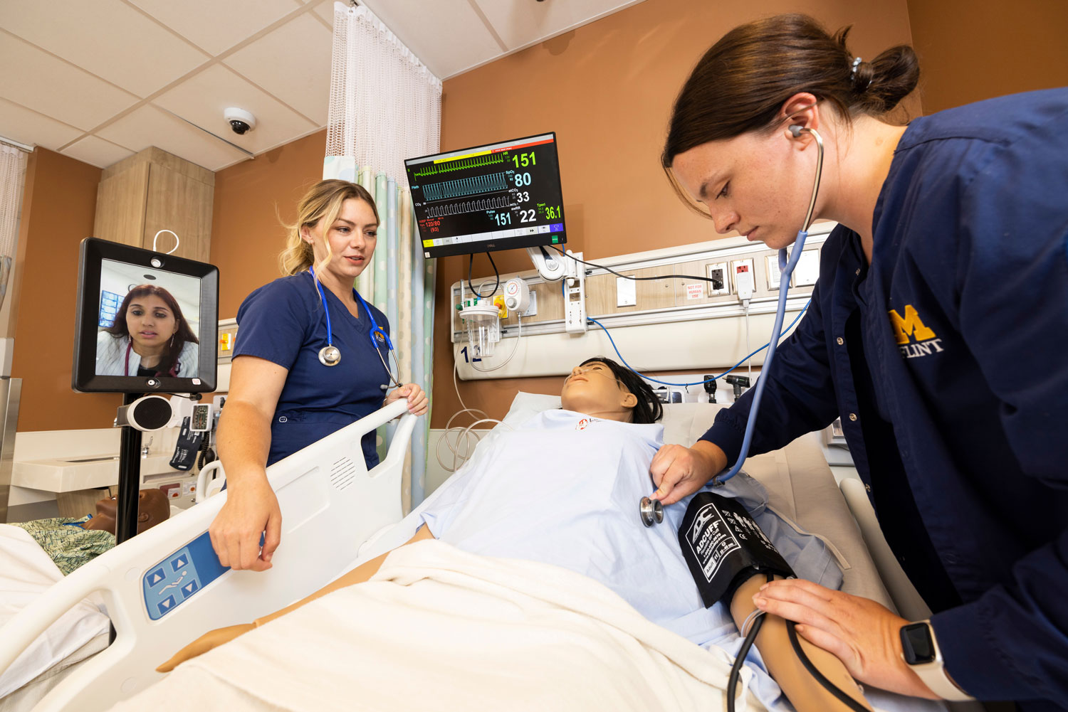 In a clinical simulation room, two nursing students, wearing U-M Flint uniforms, perform a medical checkup on a patient mannequin. One uses a stethoscope and blood pressure cuff, while a third person appears on a video screen mounted on a mobile stand.