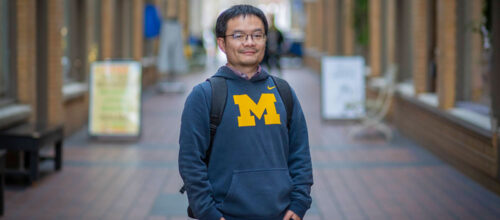 Man in a University of Michigan hoodie stands in a brick corridor, hands in pockets, looking at the camera and smiling.