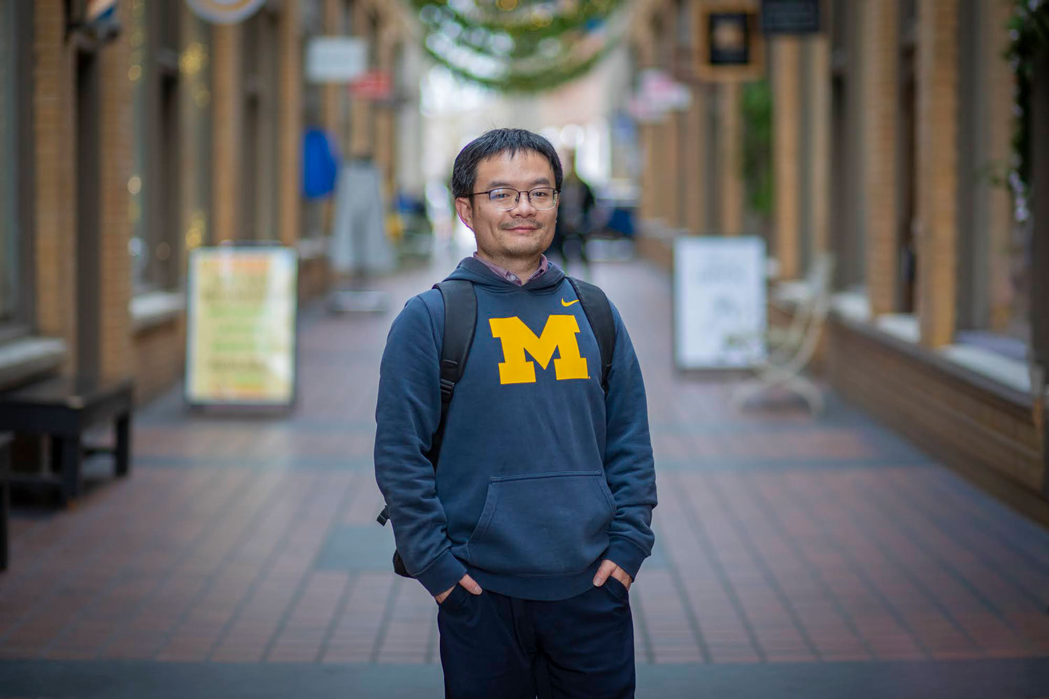 Man in a University of Michigan hoodie stands in a brick corridor, hands in pockets, looking at the camera and smiling.