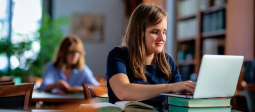 A young woman with long brown hair is seated at a green table in a library, typing on a laptop that rests atop a small stack of hardcover books. In the softly lit background, another woman with blonde hair and glasses is also reading at a separate table.