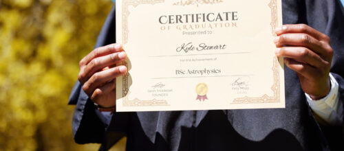 A person in a graduation gown holds up a certificate with both hands. The certificate, awarded to Kyle Stewart, recognizes the achievement of a BSc in Astrophysics.