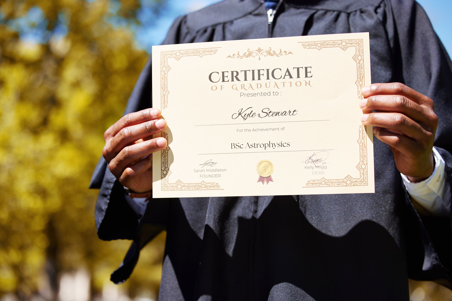 A person in a graduation gown holds up a certificate with both hands. The certificate, awarded to Kyle Stewart, recognizes the achievement of a BSc in Astrophysics.