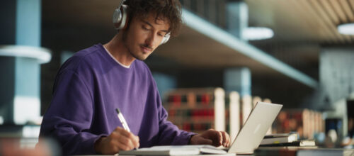 A young man wearing a purple sweatshirt and headphones is focused on writing in a notebook while using a laptop. He is seated at a modern library or study space with shelves of books in the background.