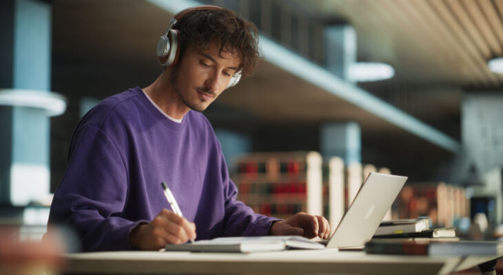 A young man wearing a purple sweatshirt and headphones is focused on writing in a notebook while using a laptop. He is seated at a modern library or study space with shelves of books in the background.