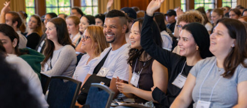 A group of diverse master's students from the University of Michigan School of Social Work sit in rows at an indoor event. Several are smiling and engaged, while a few people have raised their hands to participate.