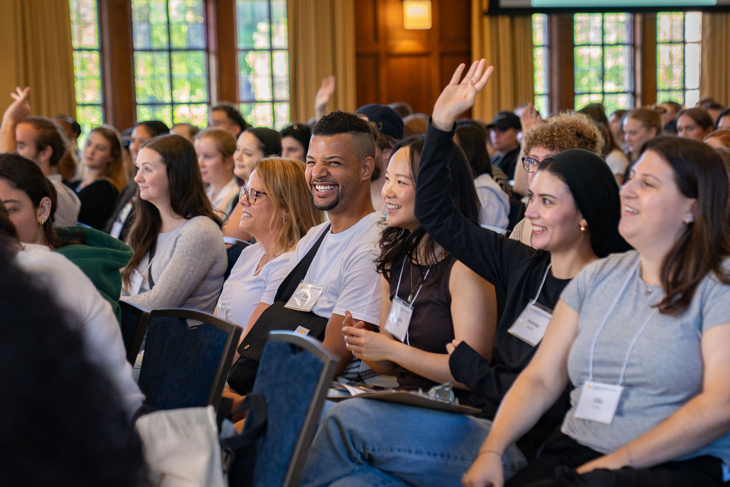 A group of diverse master's students from the University of Michigan School of Social Work sit in rows at an indoor event. Several are smiling and engaged, while a few people have raised their hands to participate.