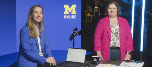 Two women stand and sit behind a desk in a studio with a blue backdrop that reads “M Online.”