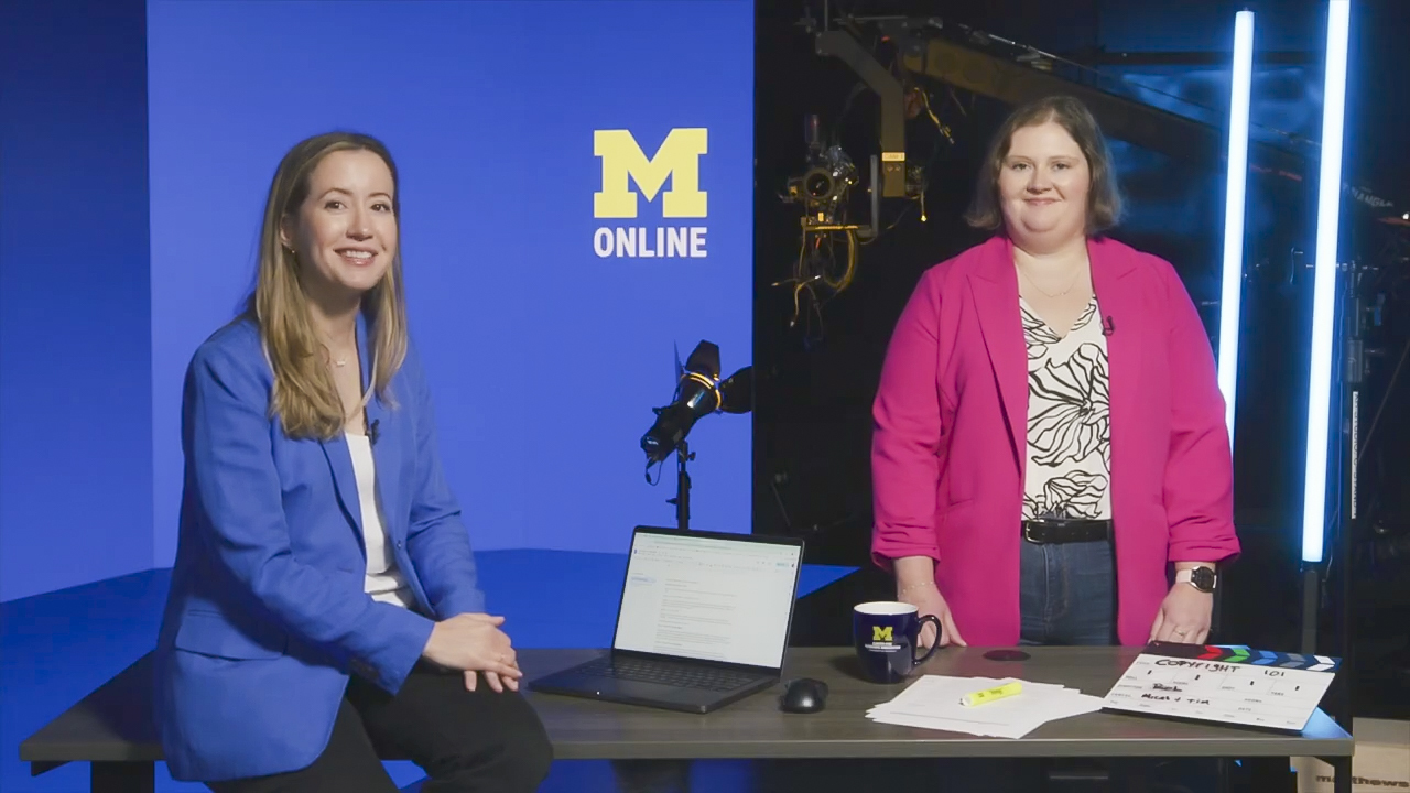 Two women stand and sit behind a desk in a studio with a blue backdrop that reads “M Online.”
