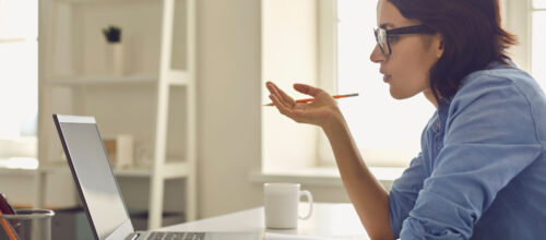 A woman in glasses and a blue shirt sits at a desk, holding a pencil near her face while looking at a laptop screen. A notebook lies open in front of her, and a white mug sits nearby in a bright, tidy room.