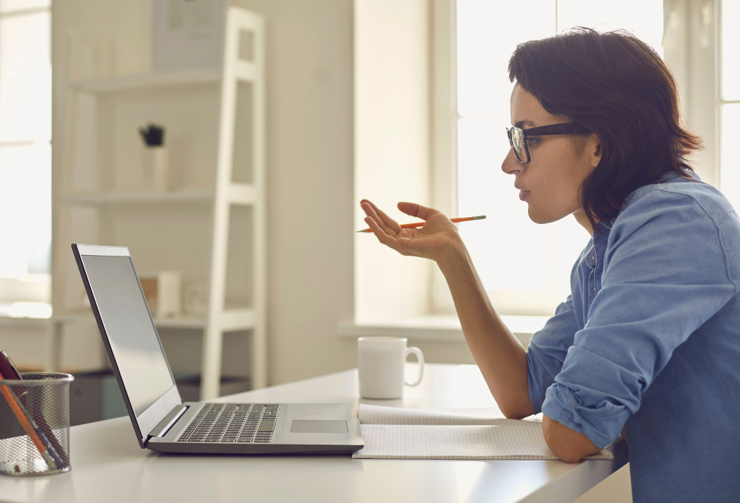 A woman in glasses and a blue shirt sits at a desk, holding a pencil near her face while looking at a laptop screen. A notebook lies open in front of her, and a white mug sits nearby in a bright, tidy room.