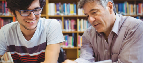 Two people sit side by side at a table in a library, looking at a laptop together, with bookshelves in the background and an engaged, collaborative posture.