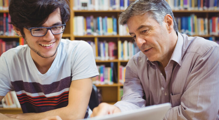 Two people sit side by side at a table in a library, looking at a laptop together, with bookshelves in the background and an engaged, collaborative posture.