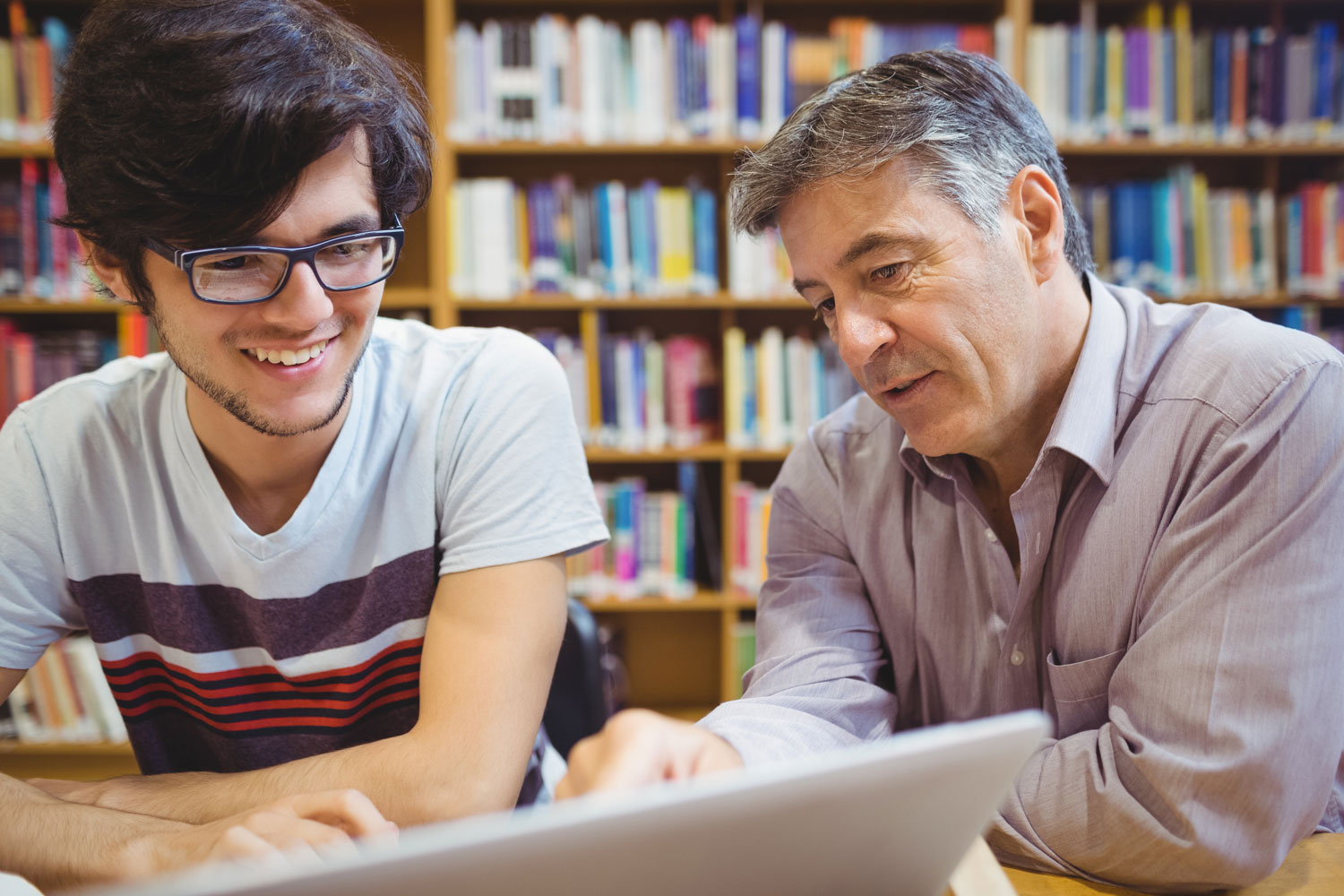 Two people sit side by side at a table in a library, looking at a laptop together, with bookshelves in the background and an engaged, collaborative posture.