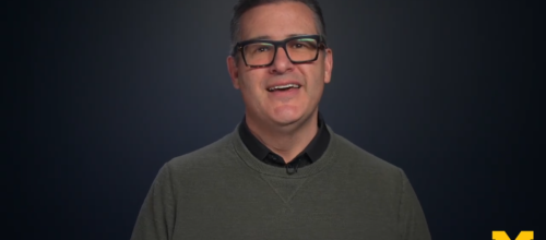 Man wearing black glasses and a green sweater smiles while facing the camera against a dark blue background with a yellow “M” logo in the lower right.