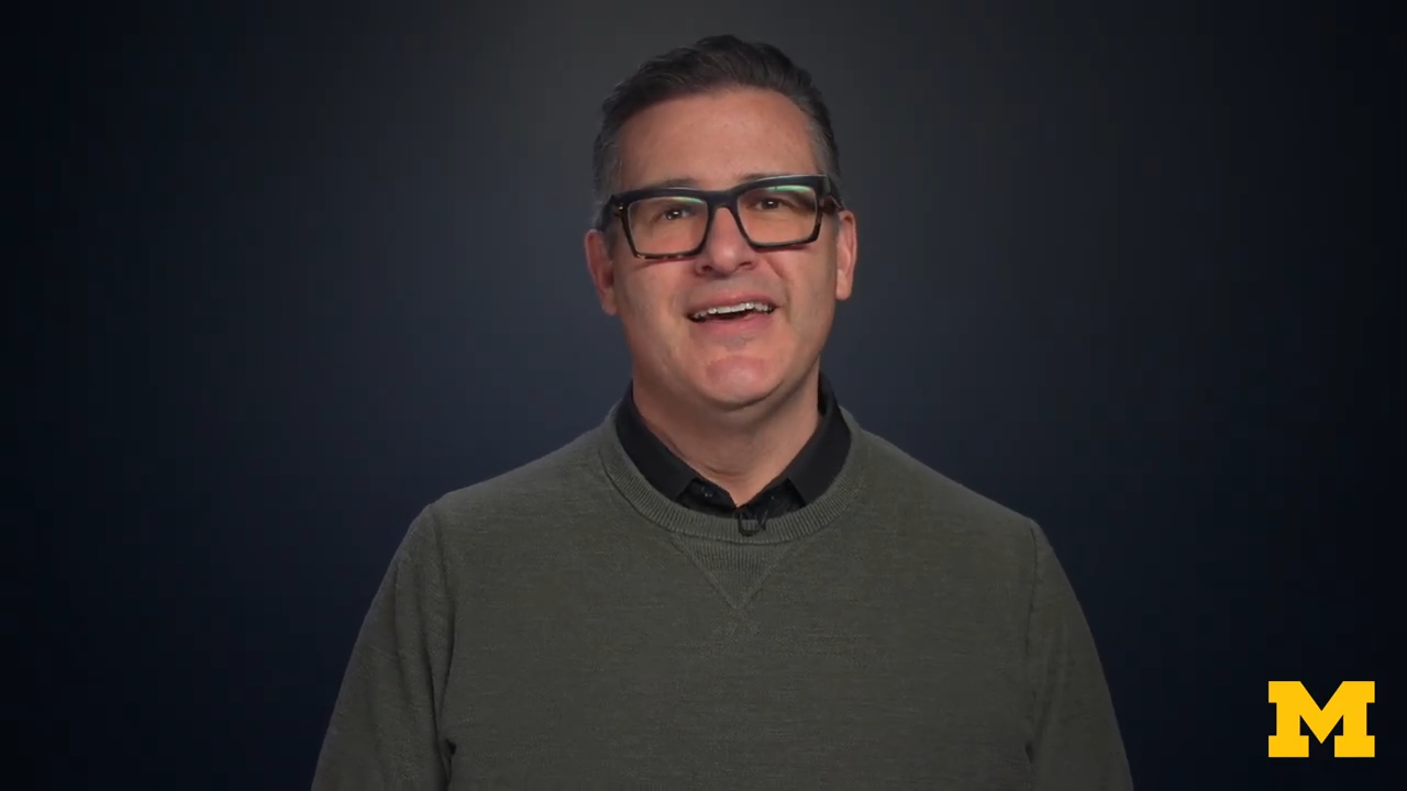 Man wearing black glasses and a green sweater smiles while facing the camera against a dark blue background with a yellow “M” logo in the lower right.