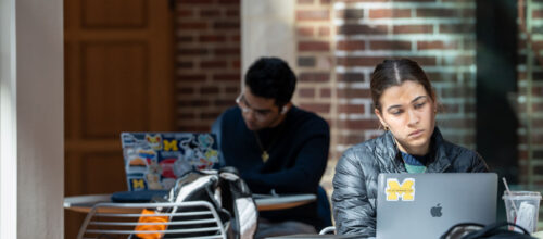 College students study at outdoor tables; woman focuses on laptop with Michigan sticker, another works behind her.