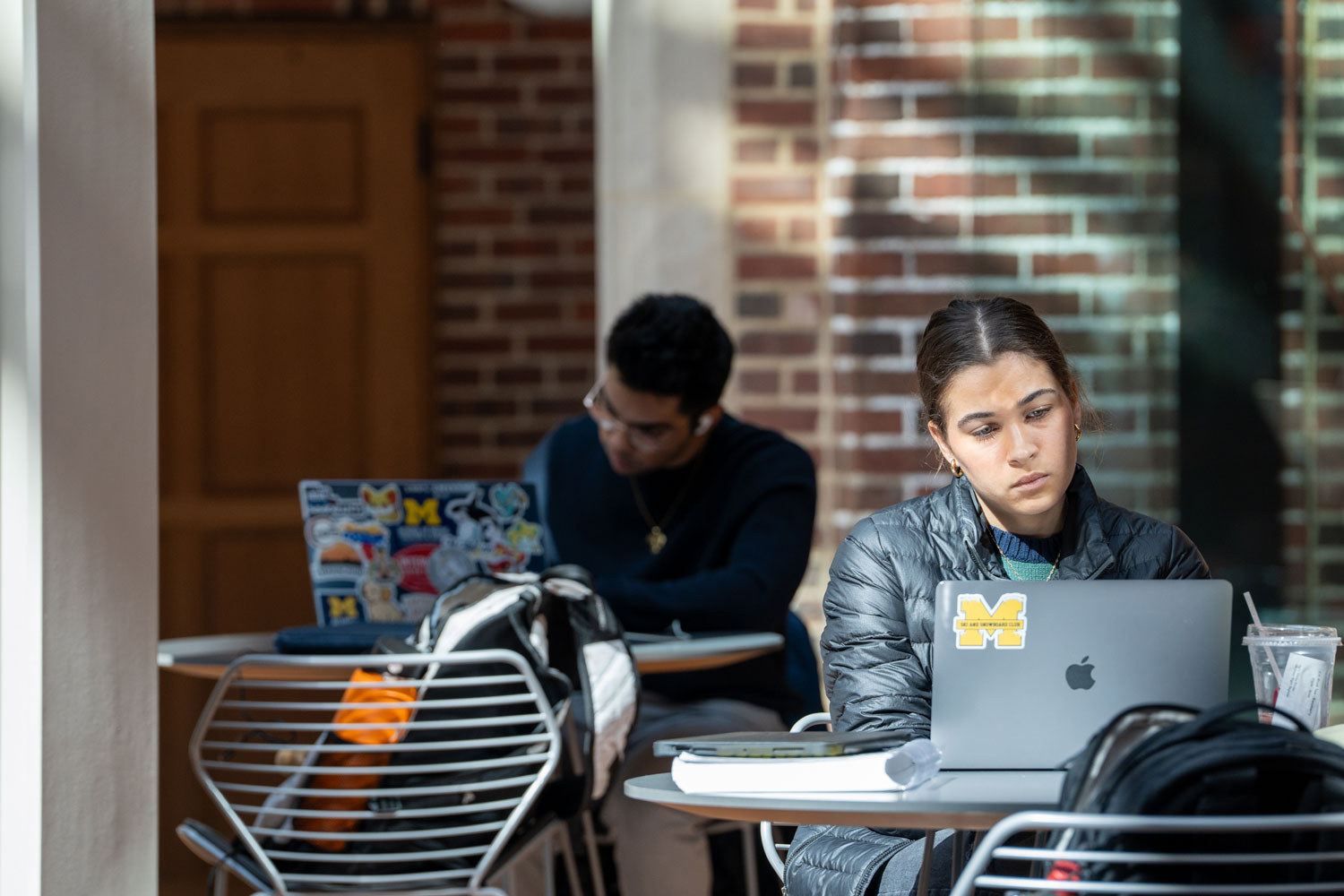 College students study at outdoor tables; woman focuses on laptop with Michigan sticker, another works behind her.