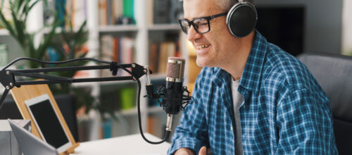 Man wearing glasses and over-ear headphones speaks into a studio microphone at a desk with a laptop and notes, suggesting he is recording a podcast or audio content.