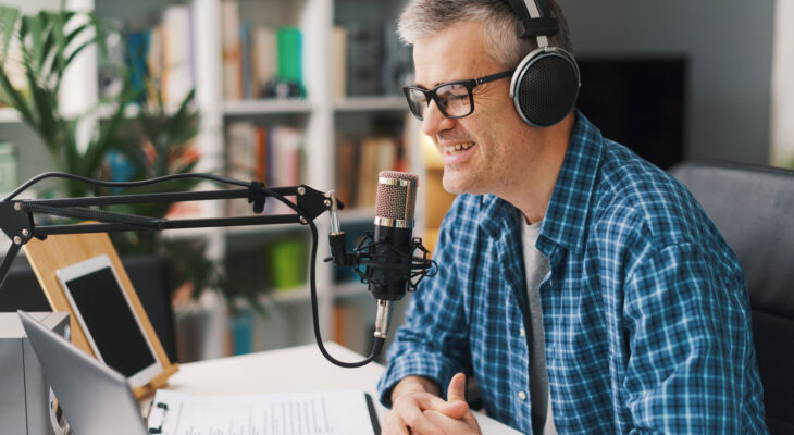 Man wearing glasses and over-ear headphones speaks into a studio microphone at a desk with a laptop and notes, suggesting he is recording a podcast or audio content.