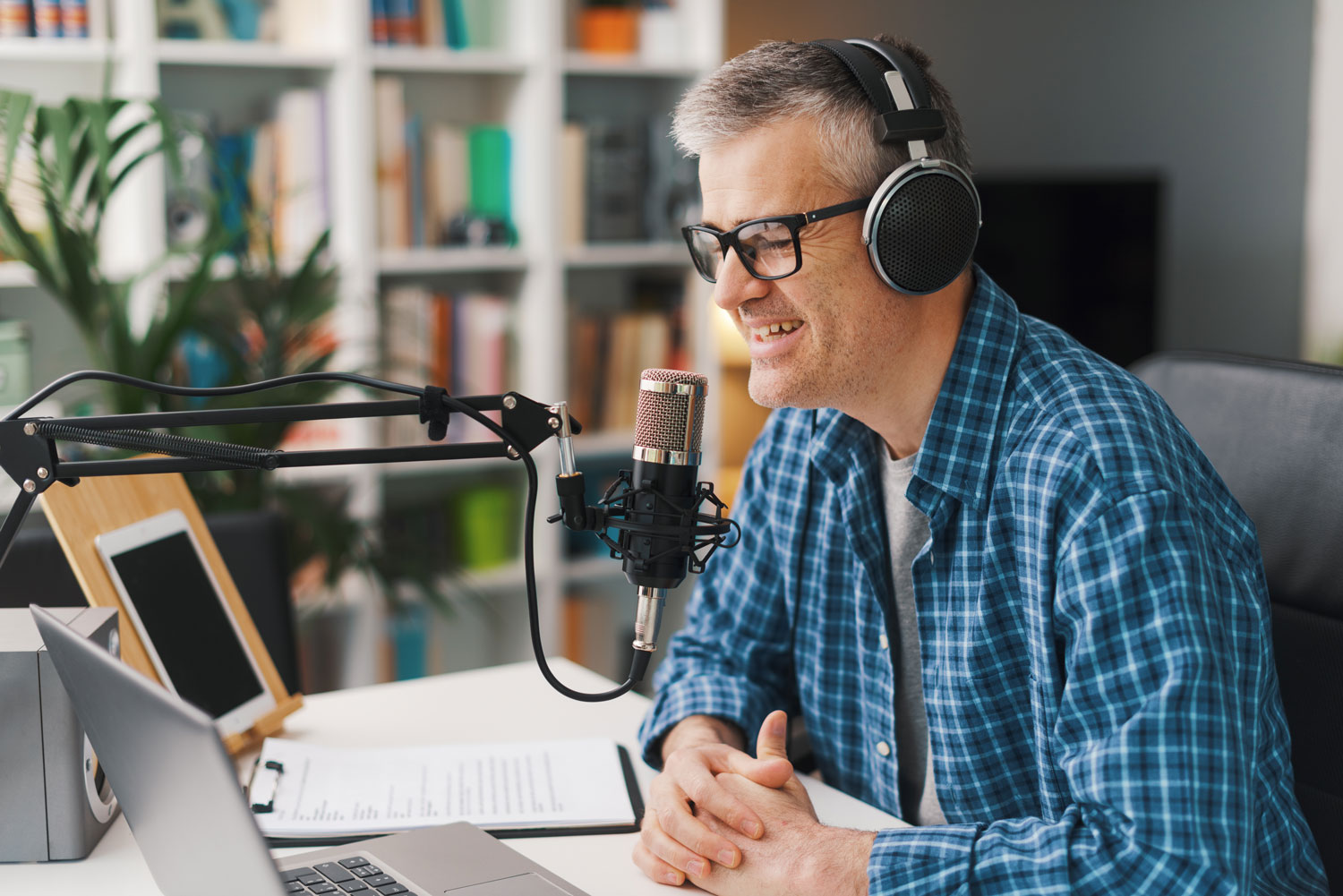 Man wearing glasses and over-ear headphones speaks into a studio microphone at a desk with a laptop and notes, suggesting he is recording a podcast or audio content.