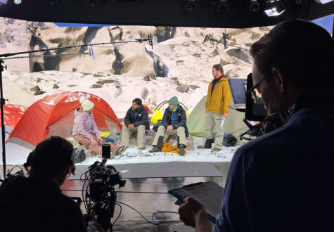 Film crew records a staged campsite scene with actors sitting by colorful tents against a snowy mountain backdrop. Lighting rigs, boom microphones, and cameras are visible around the set.