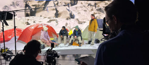 Film crew records a staged campsite scene with actors sitting by colorful tents against a snowy mountain backdrop. Lighting rigs, boom microphones, and cameras are visible around the set.