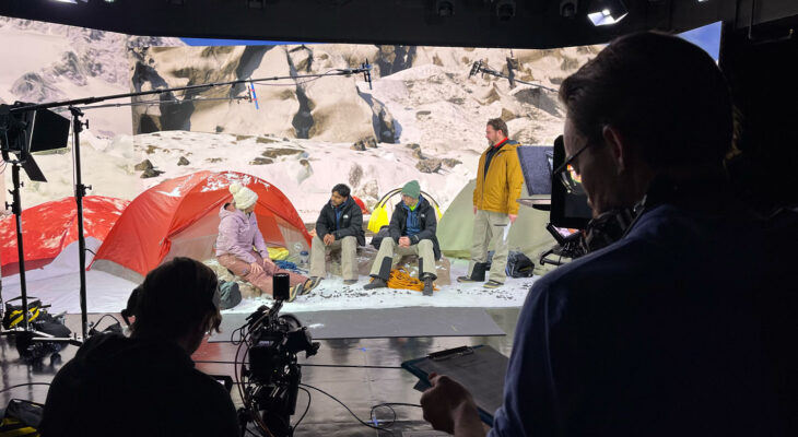 Film crew records a staged campsite scene with actors sitting by colorful tents against a snowy mountain backdrop. Lighting rigs, boom microphones, and cameras are visible around the set.