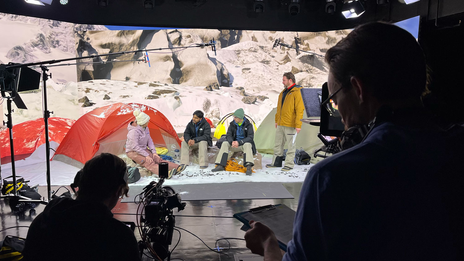 Film crew records a staged campsite scene with actors sitting by colorful tents against a snowy mountain backdrop. Lighting rigs, boom microphones, and cameras are visible around the set.