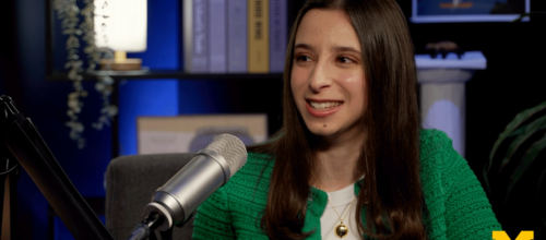 A young woman in a green jacket smiles while speaking into a microphone in a studio setting.