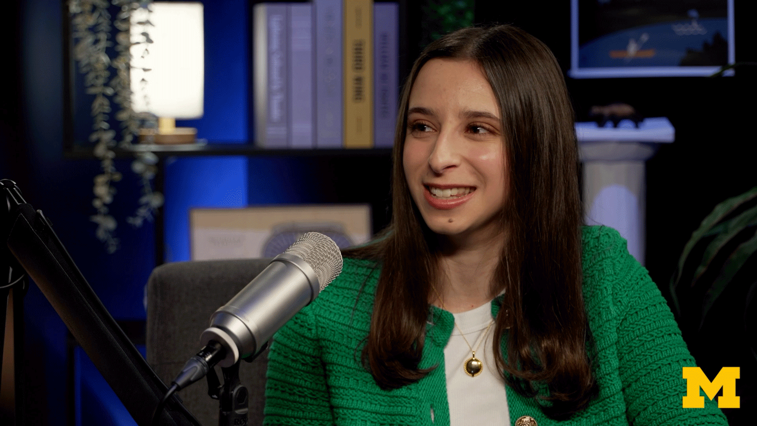 A young woman in a green jacket smiles while speaking into a microphone in a studio setting.