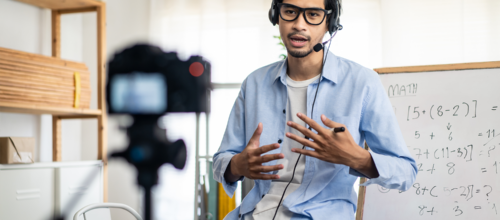 Instructor wearing a headset records a math lesson on camera, gesturing beside a whiteboard with equations.