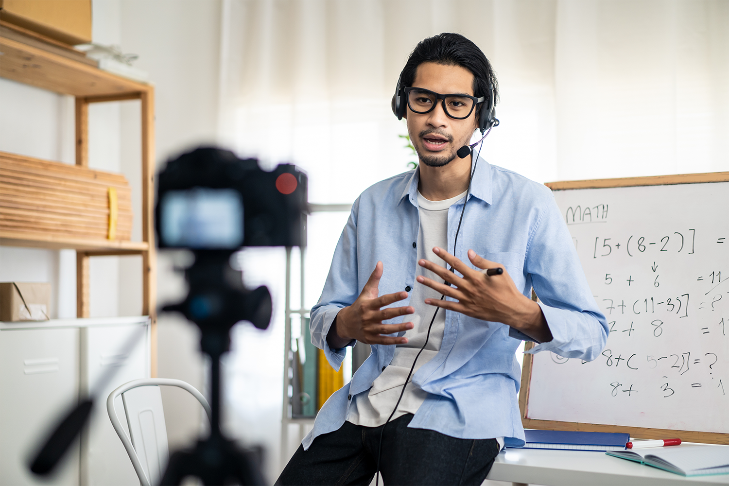 Instructor wearing a headset records a math lesson on camera, gesturing beside a whiteboard with equations.