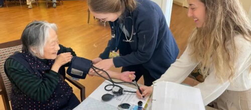 An elderly woman has her blood pressure checked by a healthcare worker while another woman assists, in a community room with people seated in the background.