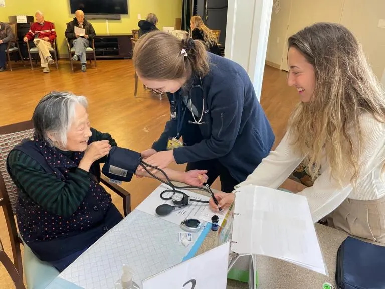 An elderly woman has her blood pressure checked by a healthcare worker while another woman assists, in a community room with people seated in the background.