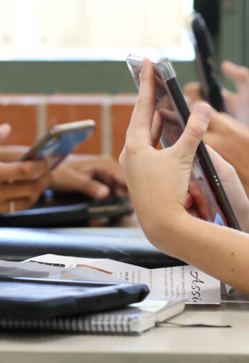 Students sitting at desks use smartphones and tablets during class while notebooks and papers rest on the tables.