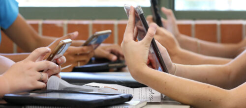 Students sitting at desks use smartphones and tablets during class while notebooks and papers rest on the tables.