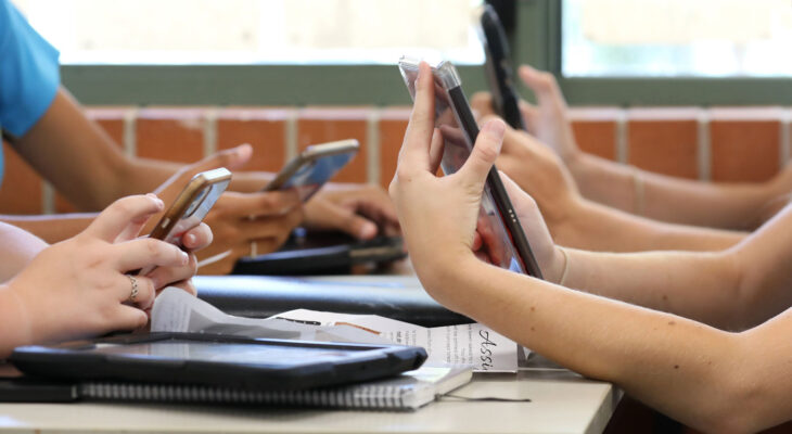 Students sitting at desks use smartphones and tablets during class while notebooks and papers rest on the tables.