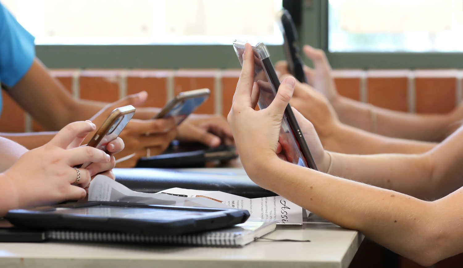 Students sitting at desks use smartphones and tablets during class while notebooks and papers rest on the tables.