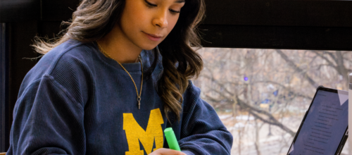 Student in a navy University of Michigan sweatshirt studies at a table by a large window, highlighting notes in a notebook while a tablet sits open nearby.