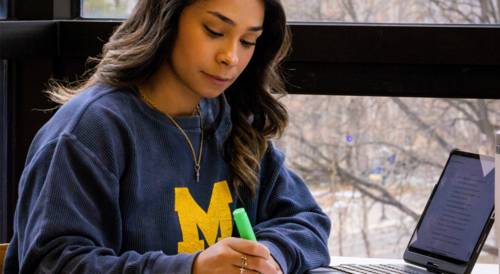 Student in a navy University of Michigan sweatshirt studies at a table by a large window, highlighting notes in a notebook while a tablet sits open nearby.
