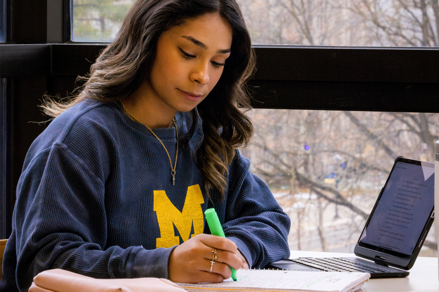Student in a navy University of Michigan sweatshirt studies at a table by a large window, highlighting notes in a notebook while a tablet sits open nearby.