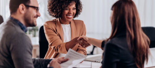 A smiling businesswoman shakes hands with a job candidate across a desk during a professional interview while another person reviews documents nearby.