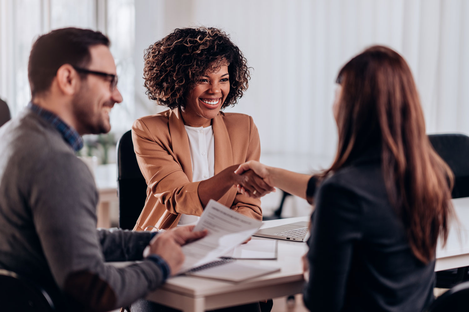 A smiling businesswoman shakes hands with a job candidate across a desk during a professional interview while another person reviews documents nearby.