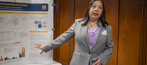 Woman in a gray blazer presents a research poster at an academic conference, gesturing toward charts and data on a display board.
