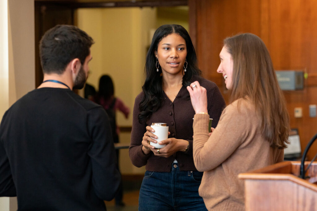 Three people stand together indoors having a casual conversation, with two holding coffee cups in a warmly lit conference or meeting space.