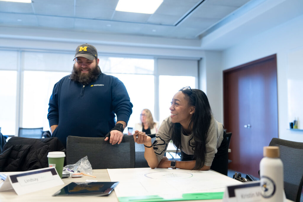 Two master's students smile and interact around a table during a workshop in a bright conference room.